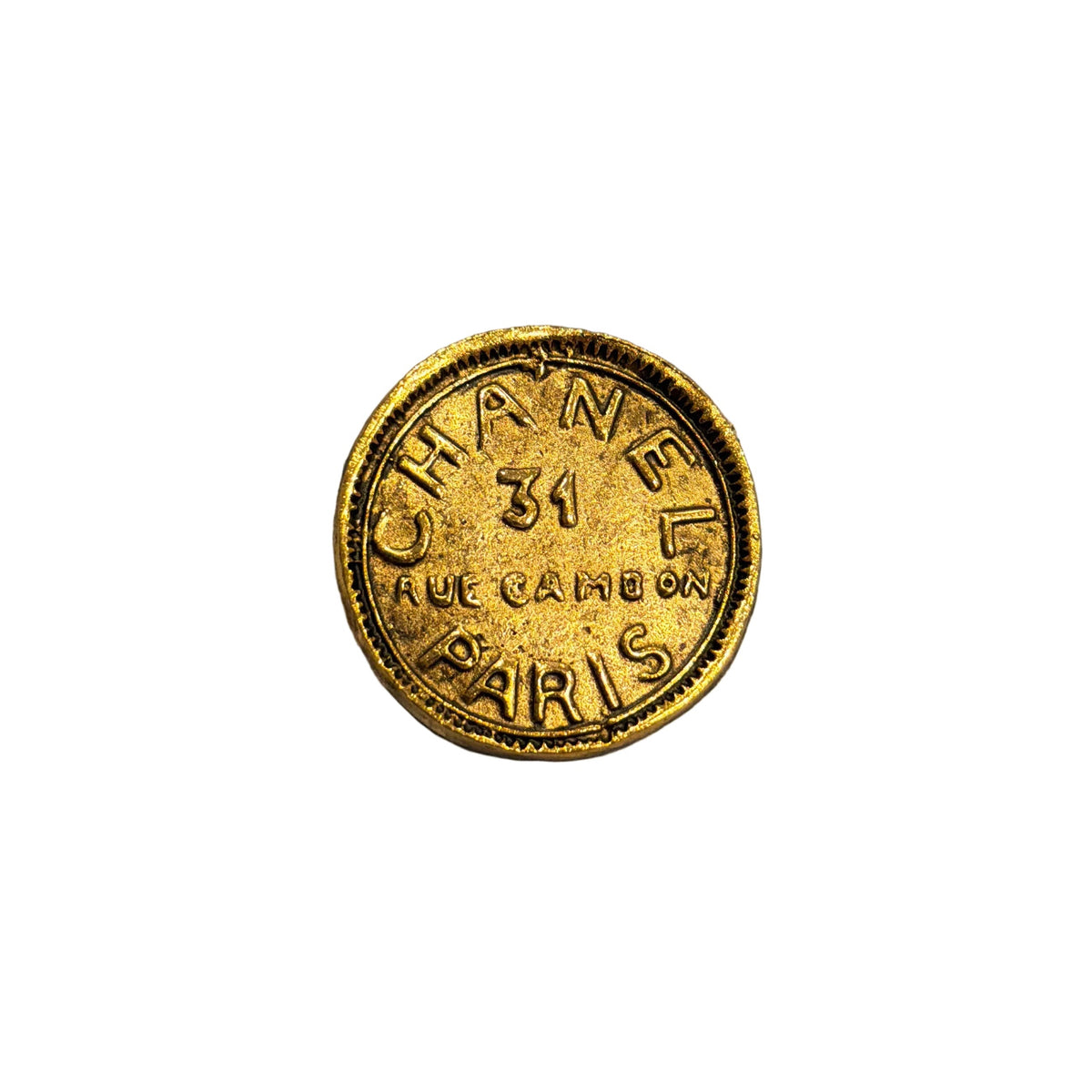 A round gold-tone Chanel pin with 'CHANEL 31 RUE CAMBRAY PARIS' embossed, resting on a gray and beige striped plush fabric background.