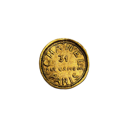 A round gold-tone Chanel pin with 'CHANEL 31 RUE CAMBRAY PARIS' embossed, resting on a gray and beige striped plush fabric background.