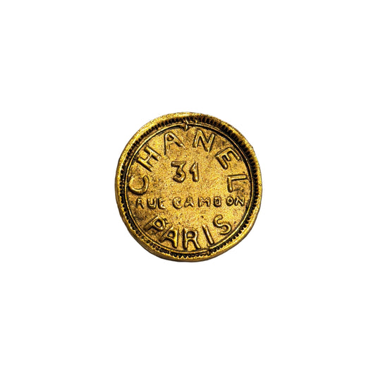 A round gold-tone Chanel pin with 'CHANEL 31 RUE CAMBRAY PARIS' embossed, resting on a gray and beige striped plush fabric background.